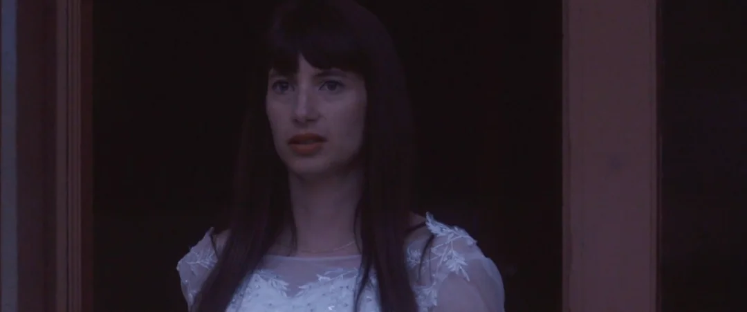 Alma : une jeune femme aux cheveux noirs en robe blanche, dans la pénombre.